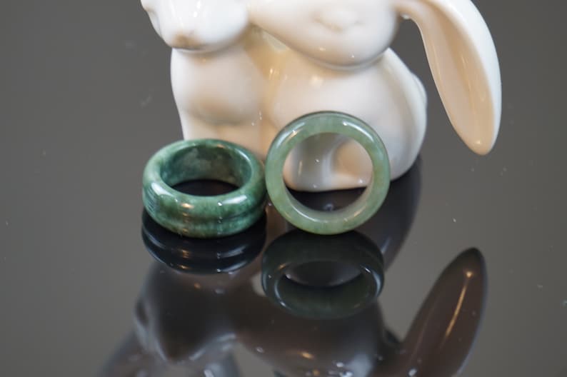 May include: Two jade green rings, one with a smooth surface and the other with a textured surface, are displayed on a white ceramic rabbit figurine.