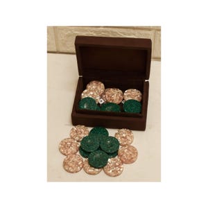 Op de afbeelding: Een bruine fluwelen doos met een open deksel, gevuld met beige en groene spelstukken. Sommige stukken zijn buiten de doos gerangschikt. Eén dobbelsteen is zichtbaar. De spelstukken zijn rond en getextureerd.