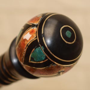 Puede incluir: Primer plano de un pomo decorativo de madera negra con dise&ntilde;os incrustados. El pomo presenta un c&iacute;rculo central turquesa, detalles dorados y secciones de mosaico naranja, blanco y turquesa. El mango tiene rayas marrones oscuras y claras alternas.