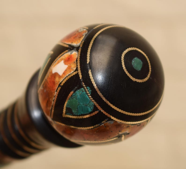 Puede incluir: Primer plano de un pomo decorativo de madera negra con dise&ntilde;os incrustados. El pomo presenta un c&iacute;rculo central turquesa, detalles dorados y secciones de mosaico naranja, blanco y turquesa. El mango tiene rayas marrones oscuras y claras alternas.