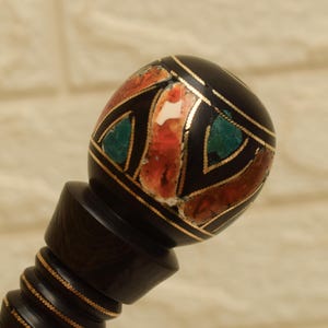 Puede incluir: Un tap&oacute;n de botella decorativo, marr&oacute;n oscuro, con una parte superior con incrustaciones. La parte superior redonda presenta un dise&ntilde;o geom&eacute;trico con detalles de piedra roja, verde y blanca, delineados con metal dorado. El tap&oacute;n tiene un tallo detallado y acanalado.