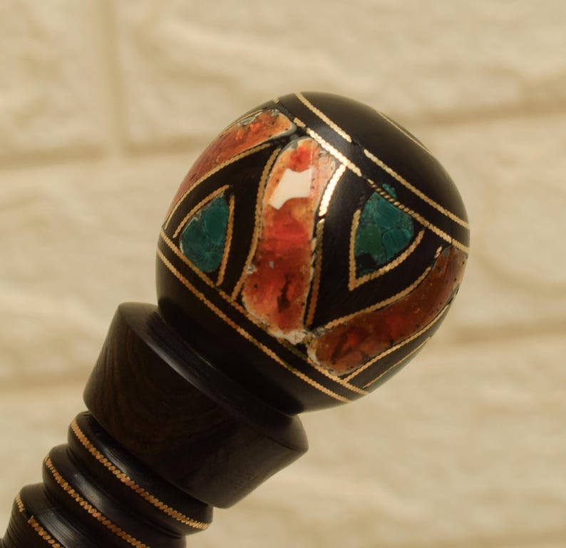 Puede incluir: Un tap&oacute;n de botella decorativo, marr&oacute;n oscuro, con una parte superior con incrustaciones. La parte superior redonda presenta un dise&ntilde;o geom&eacute;trico con detalles de piedra roja, verde y blanca, delineados con metal dorado. El tap&oacute;n tiene un tallo detallado y acanalado.