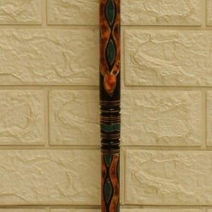 Puede incluir: Un bast&oacute;n decorativo de madera con base negra y complejos dise&ntilde;os en naranja y verde azulado. El dise&ntilde;o presenta un patr&oacute;n repetitivo de formas interconectadas. El bast&oacute;n est&aacute; sobre una pared de ladrillos texturizada de color blanquecino.