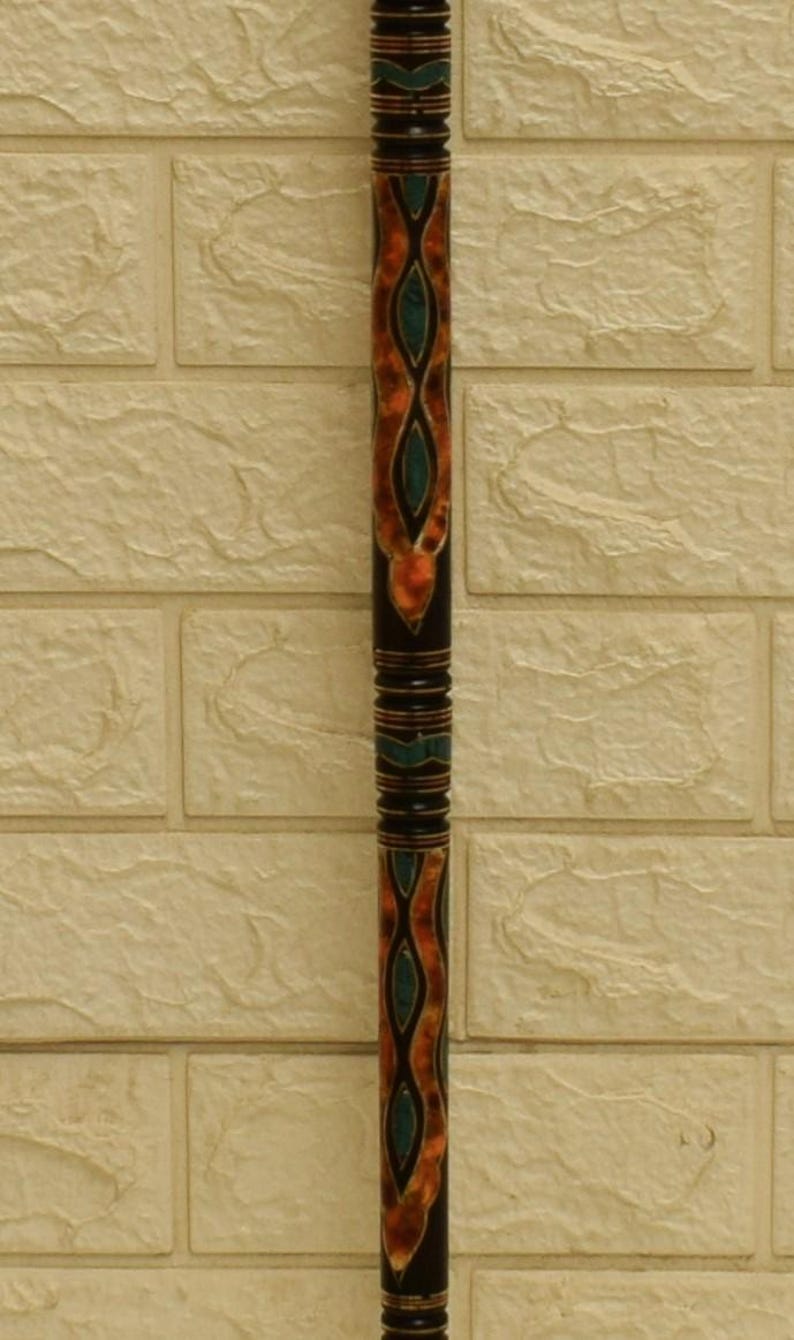 Puede incluir: Un bast&oacute;n decorativo de madera con base negra y complejos dise&ntilde;os en naranja y verde azulado. El dise&ntilde;o presenta un patr&oacute;n repetitivo de formas interconectadas. El bast&oacute;n est&aacute; sobre una pared de ladrillos texturizada de color blanquecino.
