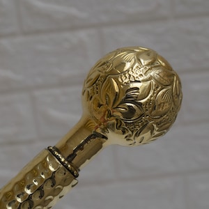 May include: A gold-colored walking stick with a round, ornate handle featuring a floral design. The handle is made of metal and has a textured surface.