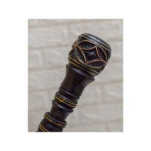 Op de afbeelding: Een donkerbruine houten wandelstok met ingewikkelde gravures en gouden en koperen accenten. De bovenkant heeft een gedetailleerd bloemmotief, terwijl de schacht een geribbeld patroon heeft. De handgreep is ongeveer 15 cm lang.