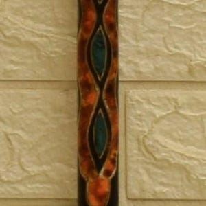 Puede incluir: Un taco de billar decorativo de madera con acabado marr&oacute;n oscuro. El eje presenta un intrincado dise&ntilde;o de formas naranjas y verde azulado. El taco de billar est&aacute; contra una pared texturizada de color blanquecino.