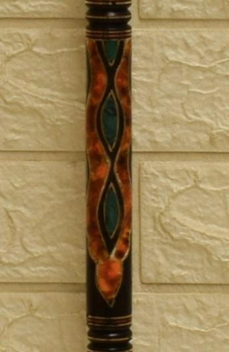 Puede incluir: Un taco de billar decorativo de madera con acabado marr&oacute;n oscuro. El eje presenta un intrincado dise&ntilde;o de formas naranjas y verde azulado. El taco de billar est&aacute; contra una pared texturizada de color blanquecino.