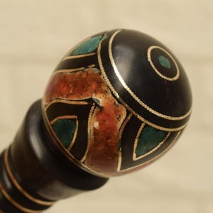 Puede incluir: Un mango decorativo, marr&oacute;n oscuro, con una parte superior redondeada y negra. La parte superior est&aacute; adornada con adornos dorados e incrustaciones de piedras turquesas y rojo anaranjado. El mango tiene una serie de anillos marrones oscuros.