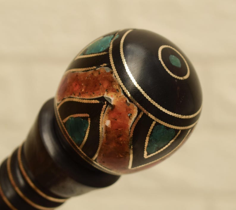 Puede incluir: Un mango decorativo, marr&oacute;n oscuro, con una parte superior redondeada y negra. La parte superior est&aacute; adornada con adornos dorados e incrustaciones de piedras turquesas y rojo anaranjado. El mango tiene una serie de anillos marrones oscuros.