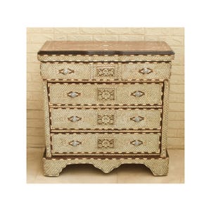 May include: A cream-coloured chest of drawers with four drawers, featuring detailed inlaid patterns and diamond-shaped accents. The top and edges are a darker brown, creating a contrast. The chest has an ornate design, suitable for a bedroom or living space.