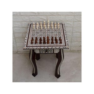 May include: A decorative chess table with a dark wood frame and inlaid mother-of-pearl details. The chess board is set with light and dark chess pieces. The table has curved legs and a small drawer.