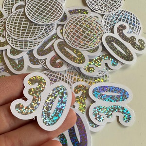 May include: A selection of holographic stickers with a white border. The stickers display the number "30" in a script font and disco ball designs. The stickers have a glittery, iridescent finish, reflecting multiple colours.