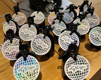 May include: Small black bottles with black caps and tags that read "#G30rgia". The tags are white with a disco ball design. The bottles are tied with black ribbon. A black cake with disco ball decorations is in the background.