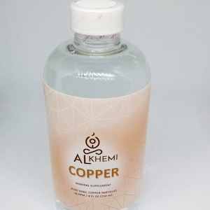 Puede incluir: Una botella de vidrio transparente con tapa blanca y etiqueta color melocotón. La etiqueta dice "ALKHEMI COPPER MINERAL SUPPLEMENT" y enumera el contenido como "PURE IONIC COPPER PARTICLES 33 PPM / 250 ml". La botella está llena de líquido transparente.