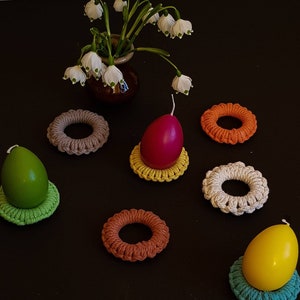 Puede incluir: Un conjunto de seis porta huevos de ganchillo en varios colores, incluyendo verde, amarillo, naranja, beige, blanco y naranja. Cada porta huevos presenta un diseño circular con una abertura central para sostener un huevo. Los porta huevos se muestran sobre una superficie negra con una sola vela blanca en cada porta huevos.