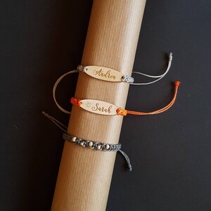 Puede incluir: Tres pulseras con etiquetas de madera con nombres. La pulsera superior es gris y dice "Andrea". La pulsera del medio es naranja y dice "Sarah". La pulsera inferior es gris con pequeñas cuentas de madera.