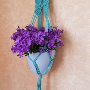 May include: A teal macrame plant hanger with a white pot and purple flowers. The hanger is hanging from a hook on a wall.