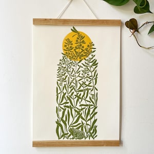 Puede incluir: Un tapiz rectangular con un estampado blanco que muestra una ilustración verde de una rana, caracoles y plantas altas bajo un sol amarillo sólido. La lámina está sujeta entre dos listones de madera y cuelga de un cordel blanco.