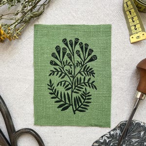 Könnte beinhalten: Ein rechteckiges Stück grüner Stoff mit einem schwarzen, im Blockdruckverfahren aufgebrachten botanischen Motiv einer blättrigen Pflanze mit kleinen, runden Blütenknospen. Der Stoff ist zusammen mit einer Metallschere, einem gelben Maßband, einem Holzbildhauerwerkzeug und einer gelben Garnrolle abgebildet.