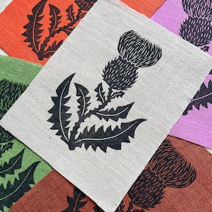 Può includere: Una collezione di ritagli di tessuto rettangolari in arancione, viola chiaro, verde, beige e ruggine, ciascuno con una stampa serigrafica nera di una pianta di cardo con un fiore testurizzato e foglie seghettate in una composizione a strati.