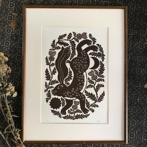 Op de afbeelding: Ingelijste kunstdruk met een bruine haas omringd door gestileerde distels en bladeren. Het kunstwerk staat tegen een witte achtergrond in een houten lijst. De print heeft een rustiek, botanisch thema.