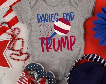 May include: A gray baby onesie with the text "Babies for Trump" in blue and red. The onesie features a red, white, and blue rattle graphic and a firework graphic. The image also includes red, white, and blue themed accessories.