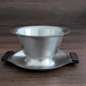 Può includere: Un set di ciotola e piattino in acciaio inossidabile. La ciotola è appoggiata su un piattino abbinato con manici in legno scuro. Il set è realizzato in metallo spazzolato ed è progettato per servire cibo o bevande.