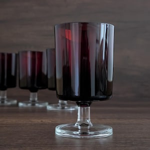 May include: A set of six burgundy-colored stemmed glasses. The glasses have a cylindrical bowl and a clear, circular base. The background is a dark brown wooden surface.