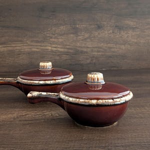 May include: Two brown ceramic soup bowls with lids. Each bowl has a handle and a white and brown rim. The lids have a small, decorative knob. The bowls are on a dark wood surface.