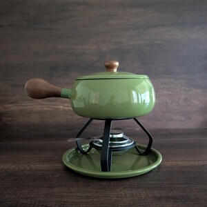 May include: A sage green fondue pot with a matching lid and a wooden handle. The pot sits on a black metal stand with a circular base and a central burner. The lid features a small wooden knob, and the entire set has a smooth, glossy finish.