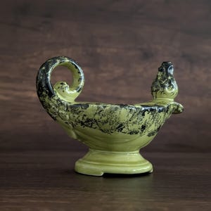 May include: A ceramic oil lamp with a green and black speckled design. The lamp has a curved handle and a spout. The base is round and the lamp is set against a dark wood background.