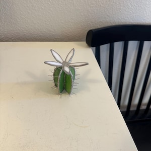 May include: A handcrafted stained glass cactus sculpture. The cactus is green with silver-colored spines and a white and clear flower on top. The sculpture is on a cream-colored surface.