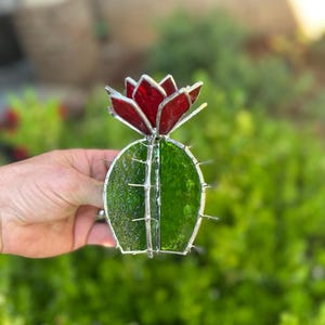 May include: A stained glass cactus sculpture with a vibrant red flower atop a green body. The cactus features a textured surface and is accented with silver solder. The piece is designed to resemble a desert plant.