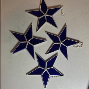 May include: Four blue stained glass star ornaments with silver-colored metal edging and attached silver chains. The stars have a five-point design and are arranged on a light gray surface.