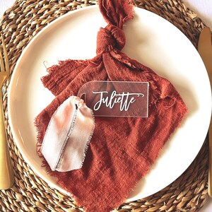 May include: A place setting with a white plate, gold fork and knife, and a rust-colored napkin. A clear acrylic name tag with the name "Juliette" is on the napkin. The plate sits on a woven placemat.