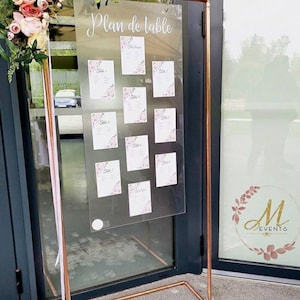 May include: A copper-colored metal wedding seating chart display. The clear acrylic board displays the words "Plan de table" and individual table assignments with floral accents. The display is decorated with flowers and ribbons.