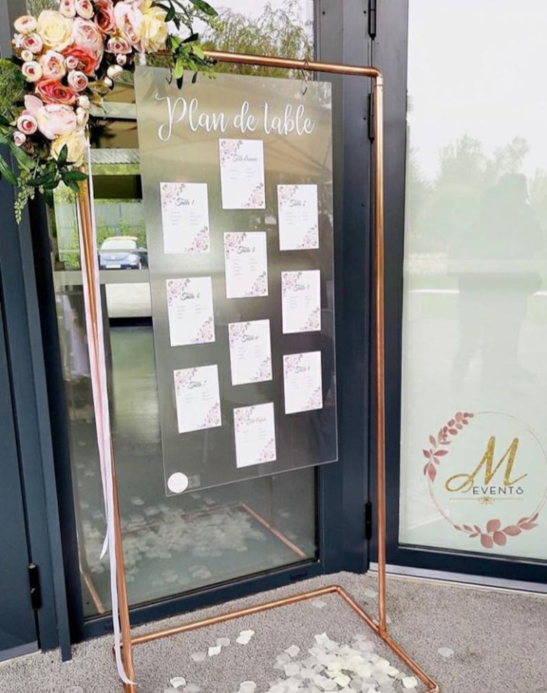 May include: A copper-colored metal wedding seating chart display. The clear acrylic board displays the words "Plan de table" and individual table assignments with floral accents. The display is decorated with flowers and ribbons.