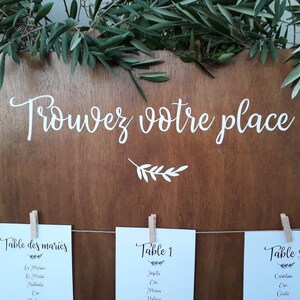 May include: A wooden sign with the text "Trouvez votre place!" in white script, adorned with greenery. Below, three cards are clipped to a string, each labeled with table numbers and names, ideal for a wedding or event.