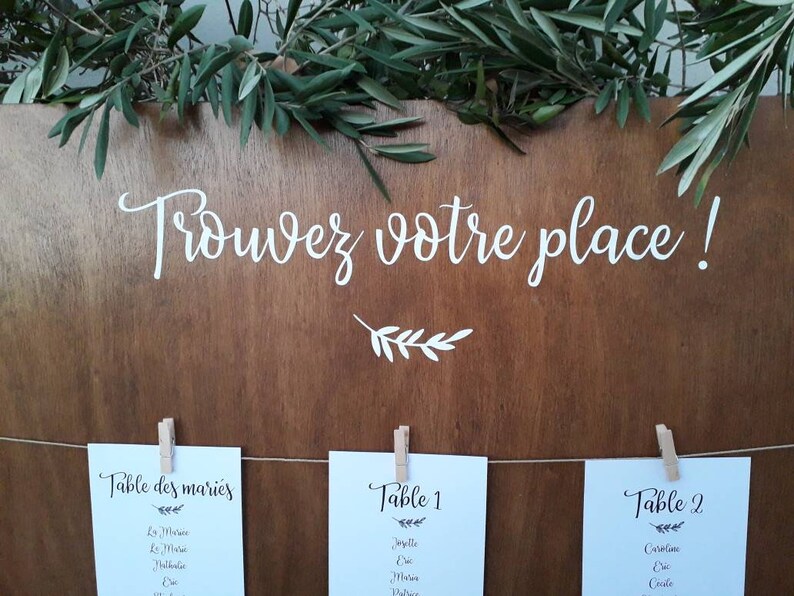 May include: A wooden sign with the text "Trouvez votre place!" in white script, adorned with greenery. Below, three cards are clipped to a string, each labeled with table numbers and names, ideal for a wedding or event.