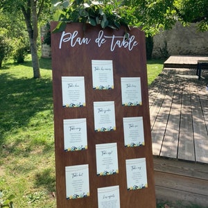 May include: A wooden wedding seating chart with the words "Plan de table" in white script. The chart displays several smaller cards with guest names and table assignments, decorated with floral accents. Greenery is placed at the top.