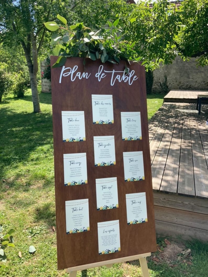 May include: A wooden wedding seating chart with the words "Plan de table" in white script. The chart displays several smaller cards with guest names and table assignments, decorated with floral accents. Greenery is placed at the top.