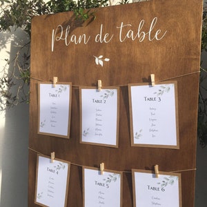 May include: A wooden wedding seating chart with the words "Plan de table" in white script. Six cards are clipped to the board with names listed under the table numbers. Greenery accents the cards and the top of the board.