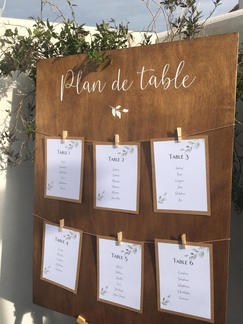 May include: A wooden wedding seating chart with the words "Plan de table" in white script. Six cards are clipped to the board with names listed under the table numbers. Greenery accents the cards and the top of the board.