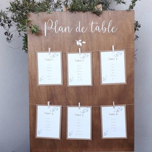 May include: A wooden wedding seating chart with the words "Plan de table" in white script. Six cards are clipped to the board with clothespins, each listing table numbers and names. Greenery decorates the top of the board.
