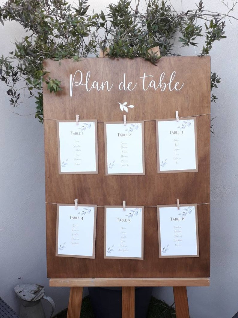 May include: A wooden wedding seating chart with the words "Plan de table" in white script. Six cards are clipped to the board with clothespins, each listing table numbers and names. Greenery decorates the top of the board.