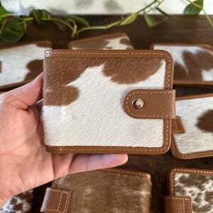 May include: A collection of small, bi-fold wallets. The wallets are made of cowhide with a brown and white pattern. The edges and the snap closure are brown. The wallets are displayed on a wooden surface.