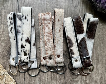 May include: A collection of leather keychains with cowhide patterns in black and white, brown and white, and tan and white. Each keychain has a metal ring and a leather loop for easy attachment. The keychains are arranged on a wooden surface.