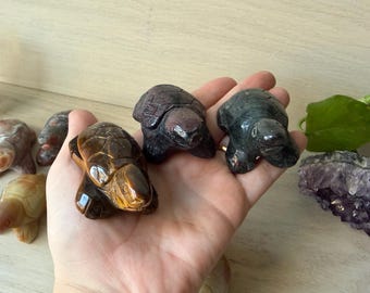 May include: A hand holds three carved stone turtle figurines in shades of brown, black, and green. Other turtle figurines in various colors are visible in the background. A cluster of purple amethyst crystals and a green leaf are also present.