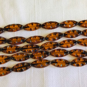 May include: Rows of amber-colored, oval-shaped beads with dark brown and silver accents. The beads are strung together, showcasing a repeating pattern. The beads are likely for jewelry making or crafting.